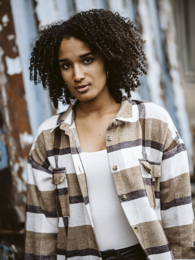 Nahaufnahme von Model Clara mit Locken und kariertem Hemd vor rustikalem Hintergrund