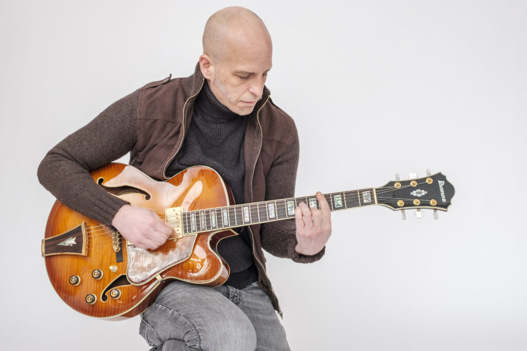 Sitzender Gitarrist beim Spielen einer orangefarbenen halbakustischen E-Gitarre vor weißem Hintergrund.