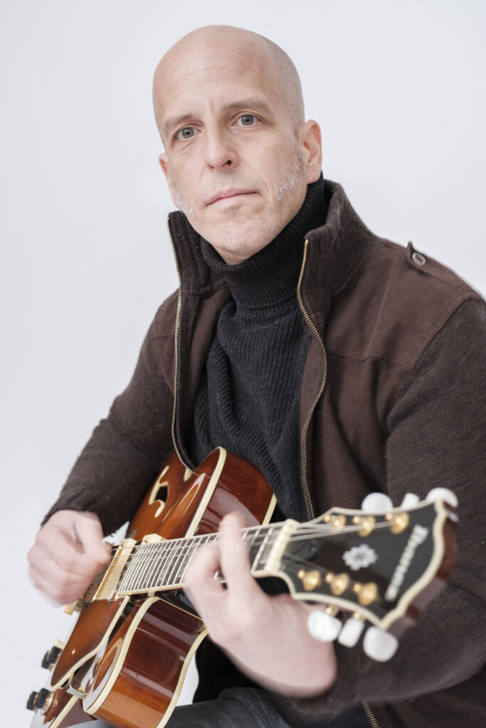 Künstlerporträt eines Gitarristen beim Spielen einer halbakustischen E-Gitarre vor hellem Hintergrund.