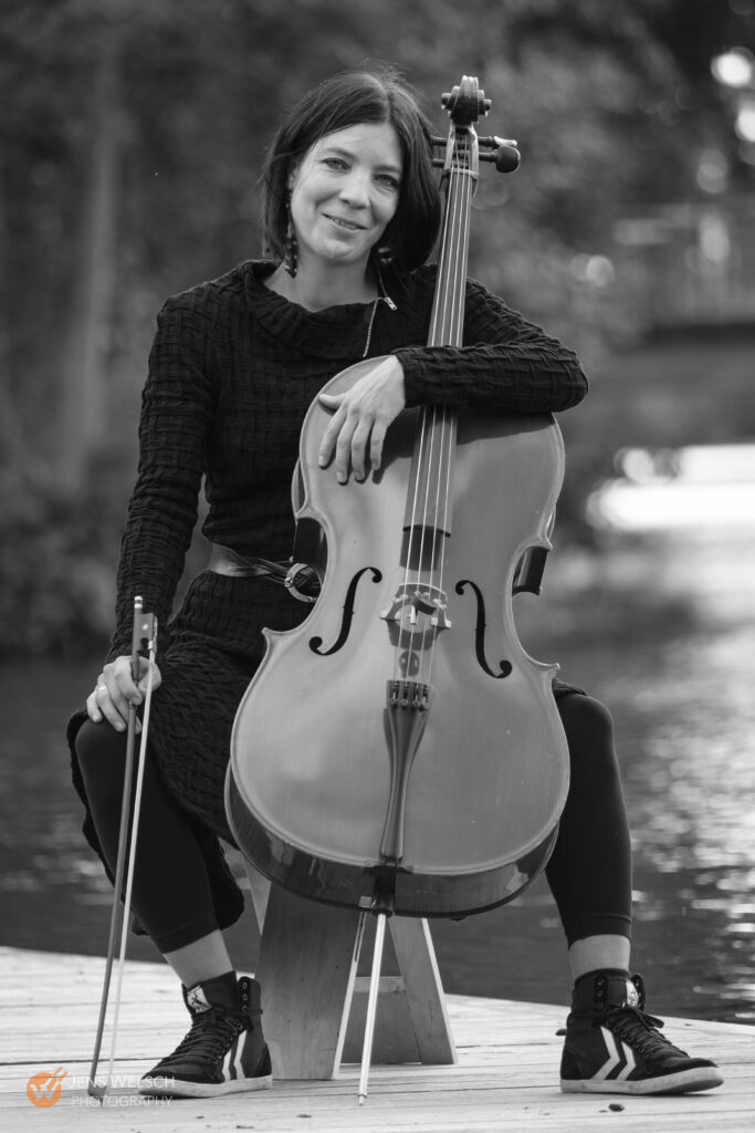 Schwarz-Weiß-Künstlerporträt einer Cellistin mit Cello und Bogen im Freien am Wasser.