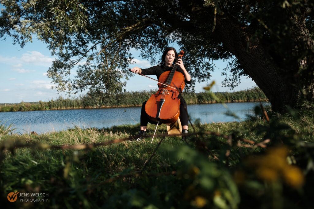 Cellistin spielt ihr Cello unter einem großen Baum am Wasser in einer natürlichen Landschaft.