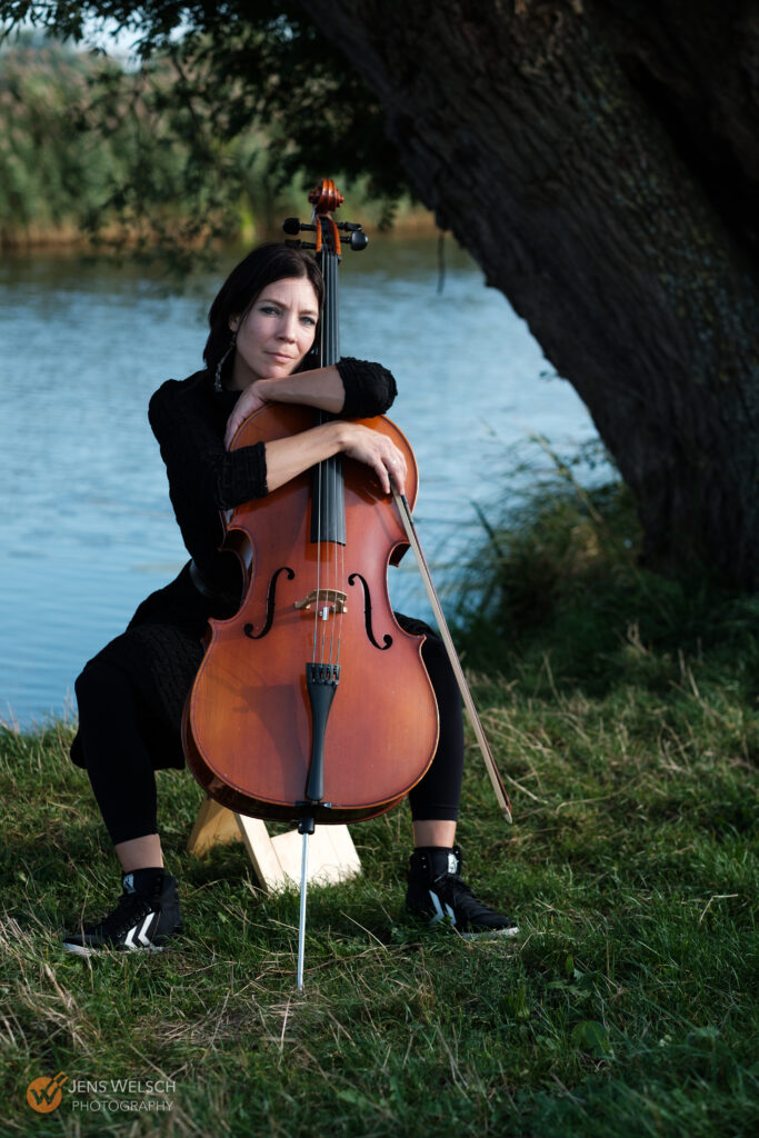 Cellistin mit Cello und Bogen unter einem großen Baum am Ufer eines Gewässers.