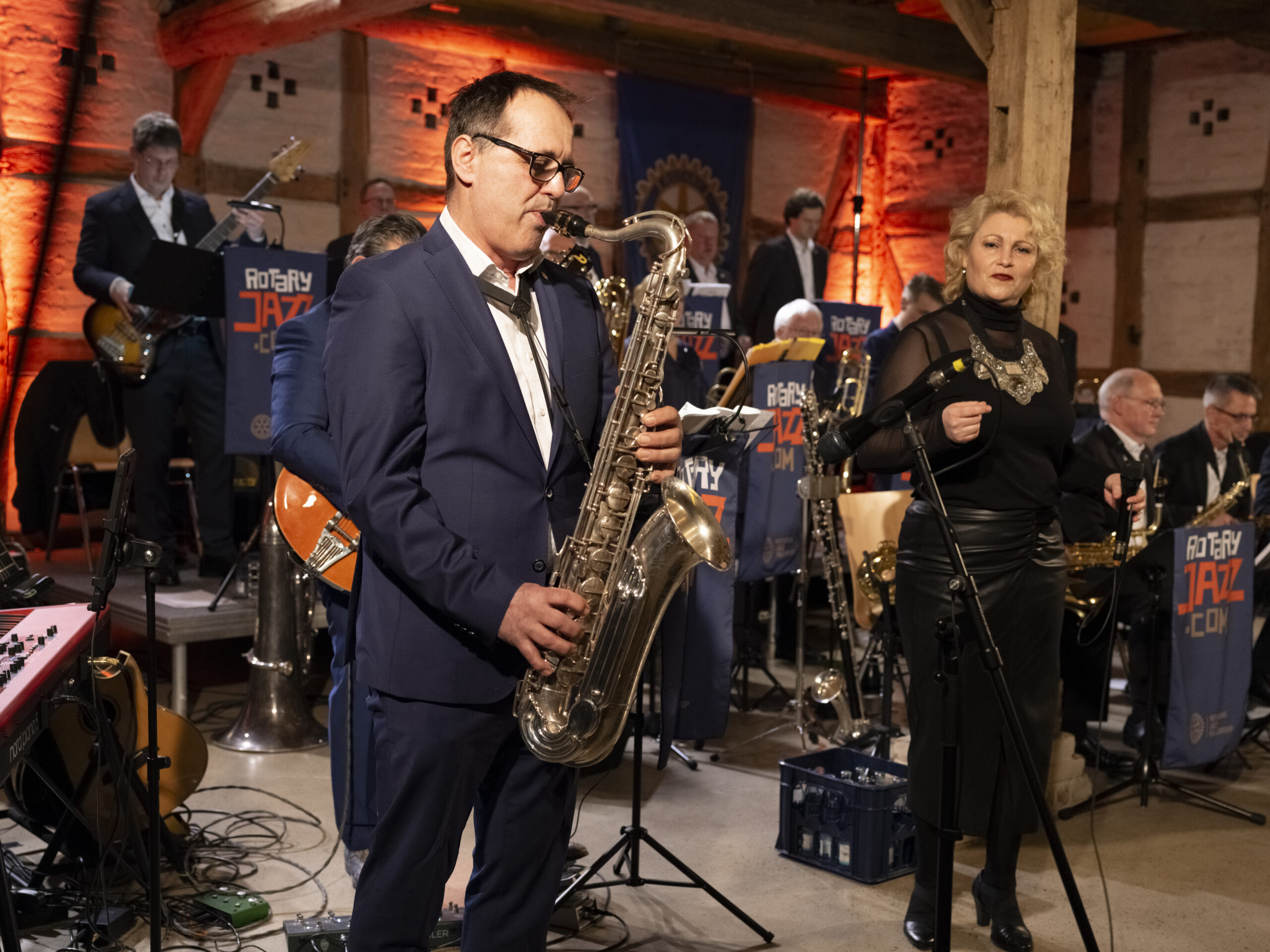 Saxophonist im Anzug spielt ein Saxophon-Solo auf der Bühne neben einer Sängerin bei rotem Bühnenlicht.
