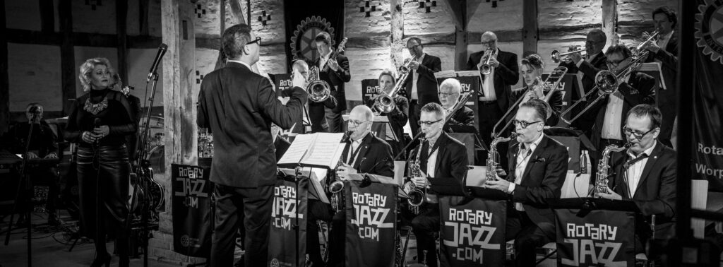 Der Dirigent Thomas Siffling leitet die Bläsersätze einer Bigband bei einer Aufführung auf der Bühne in Schwarz-Weiß.