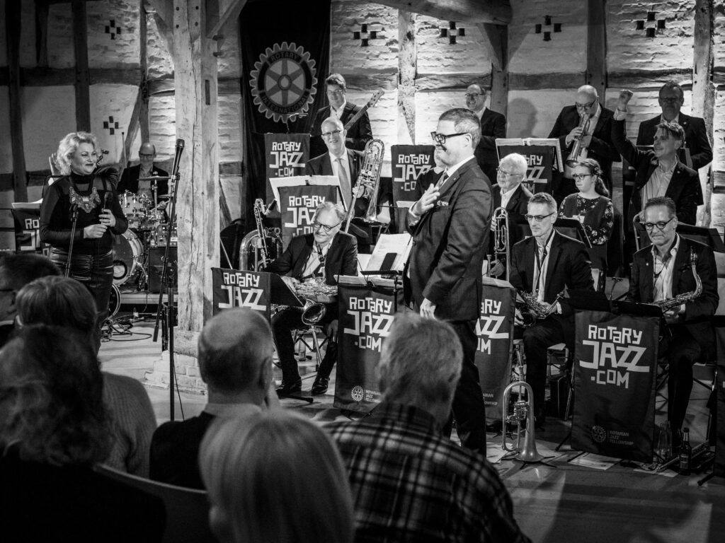 Der Bandleader spricht mit der Hand auf dem Herzen zum Publikum vor der stehenden Bigband.