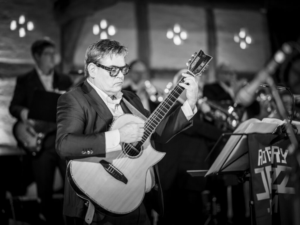 Nahaufnahme eines Gitarristen mit Brille beim Spiel einer Akustikgitarre in Schwarz-Weiß auf der Bühne.