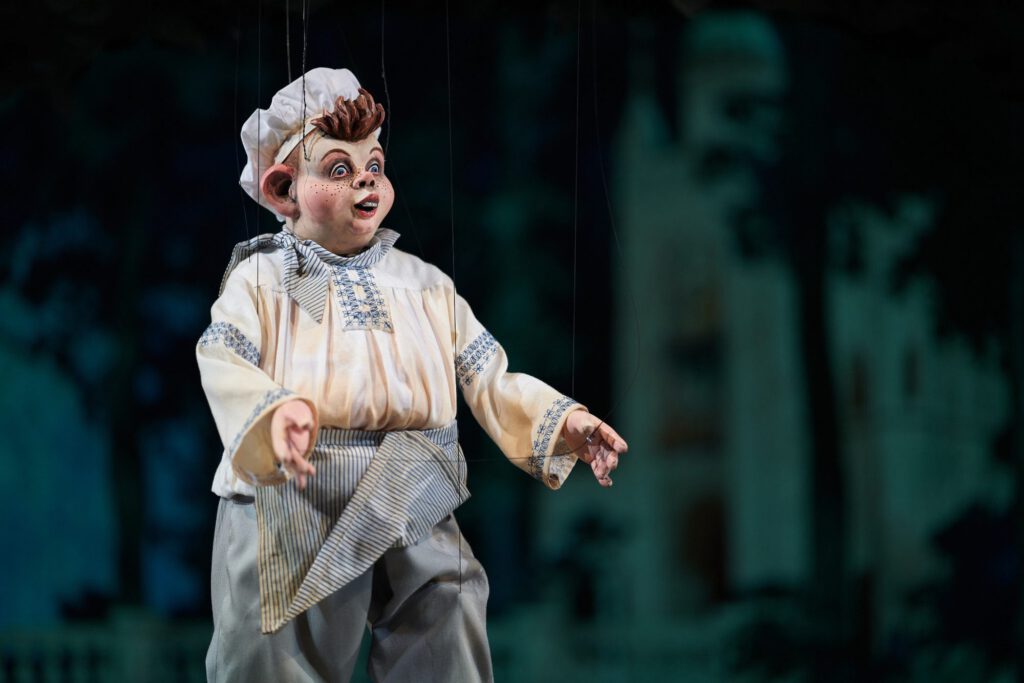 Nahaufnahme einer Marionette in heller Kochkleidung vor dunkler Bühnenkulisse.