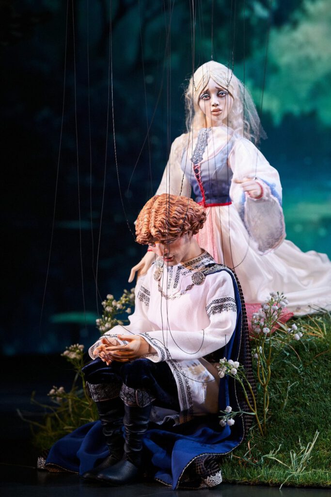 Rusalka und der Prinz - Zwei Marionetten sitzen hintereinander zwischen Gräsern und weißen Blüten auf der Bühne.