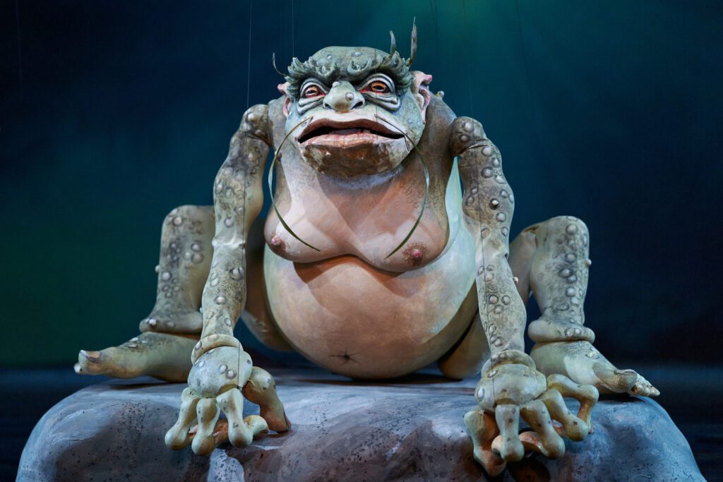 Der Wassermann - Große amphibienartige Marionette sitzt frontal auf einem Felsen vor blaugrünem Bühnenhintergrund.